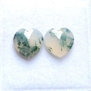 May include: Two heart-shaped moss agate gemstones with a faceted surface. The stones are translucent and off-white, featuring distinct dark green, branch-like inclusions that resemble moss patterns spread throughout the interior of each stone.