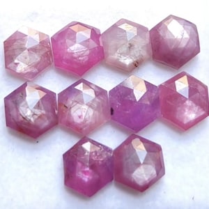 Puede incluir: Una colección de diez piedras preciosas sueltas con corte hexagonal en varios tonos de rosa y magenta. Cada piedra presenta una superficie facetada que refleja la luz, mostrando inclusiones internas y variaciones naturales de color.