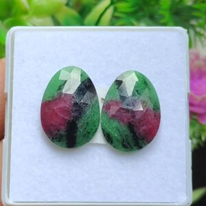 May include: Two oval gemstones with a faceted cut. The stones display a vibrant mix of colours, including deep red, forest green, and black. The gemstones are presented in a white box, highlighting their polished surfaces and unique colour patterns.
