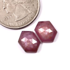 May include: Two faceted pink ruby gemstones, each with a hexagonal shape, are shown next to a US quarter for size comparison.