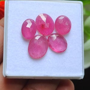 Puede incluir: Cinco gemas rosas ovaladas y facetadas se muestran en una caja blanca. Las gemas tienen una apariencia vibrante y translúcida, con estructuras internas visibles. Las facetas capturan la luz, creando un efecto brillante. Son probablemente rubíes.