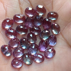 May include: A collection of faceted, oval-shaped gemstones in shades of purple, pink, and blue. The gems are scattered across a surface, showcasing their varying colors and internal structures. The stones have a polished, reflective surface.