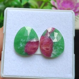 Op de afbeelding: Twee gefacetteerde edelstenen in de vorm van een druppel. De edelstenen zijn een mix van groen en rood, met de kleuren verdeeld in een patchworkpatroon. De edelstenen zijn geplaatst tegen een witte achtergrond.