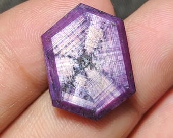 Puede incluir: Una gema facetada, hexagonal, de color púrpura intenso. La gema presenta patrones internos blancos y grises, creando una textura visual única. La piedra se sostiene en una mano, mostrando su tamaño y color.