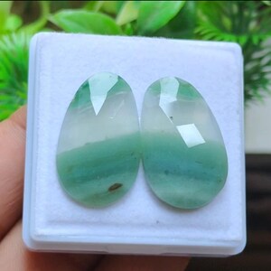 May include: Two faceted, teardrop-shaped gemstones. The stones are a mix of white and green, with a layered appearance. They are set in a white box, with green foliage in the background.