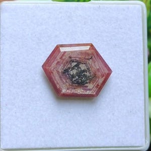 May include: A faceted, hexagonal gemstone with a reddish-brown hue. The center of the stone features a dark, textured inclusion. The gem is set against a white background, showcasing its intricate internal structure and polished facets.