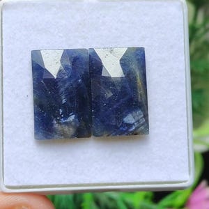Puede incluir: Dos zafiros rectangulares facetados, con tonos azul oscuro y plateado, presentados en un estuche blanco. Las piedras preciosas tienen una superficie pulida y un corte geométrico, mostrando sus variaciones de color naturales.