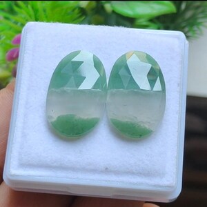 May include: Two oval gemstones with faceted surfaces, displaying a colour gradient. The top is a clear, light green, fading to white, and then a darker green, mossy base. The gems are presented in a white box.