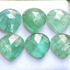 May include: Six faceted heart-shaped emerald gemstones arranged in two rows of three. Each stone displays a translucent light green hue with visible internal inclusions and a polished surface that reflects light across its geometric facets.