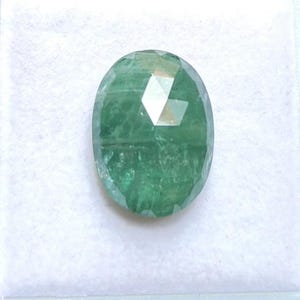 May include: An oval-shaped, faceted green gemstone with a translucent appearance and visible internal inclusions. The stone features a polished surface with multiple geometric facets that reflect light, displaying varying shades of deep and light green.