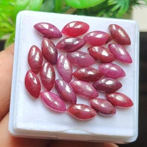 Pode incluir: Uma coleção de pedras preciosas de rubi ovais e polidas em tons de vermelho e rosa. As pedras preciosas são exibidas em um recipiente branco, mostrando sua cor natural e brilho. Perfeito para a criação de joias.