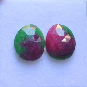 May include: Two oval-shaped gemstones with faceted surfaces. One is primarily green with a deep red center, the other is mostly red with green edges. The gems are set against a white background, showcasing their vibrant colors and cut.