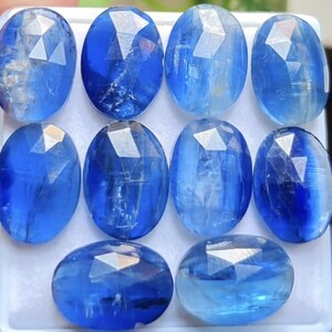 May include: A collection of ten oval-shaped, faceted blue kyanite gemstones arranged in a grid. Each stone displays a translucent quality with varying shades of deep to light blue, featuring a polished surface with geometric facets reflecting light.