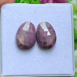 Op de afbeelding: Twee ovaalvormige, gefacetteerde edelstenen met een paarsrode tint. De edelstenen worden getoond in een witte doos, die hun gepolijste oppervlakken en interne patronen laat zien. De stenen hebben een hoge glans en worden waarschijnlijk gebruikt voor sieraden.