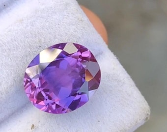 May include: An oval-cut gemstone with a vibrant purple hue, exhibiting facets that catch and reflect light. The gem is set against a white fabric background, highlighting its color and clarity. This gemstone is suitable for jewelry.