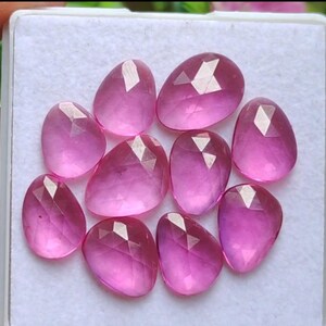 Könnte beinhalten: Eine Sammlung von neun facettierten, oval geformten rosa Edelsteinen. Die Edelsteine sind in einer weißen Box angeordnet und zeigen jeweils ein komplexes Muster interner Reflexionen. Die Steine variieren leicht im Farbton, von hell- bis tiefrosa.