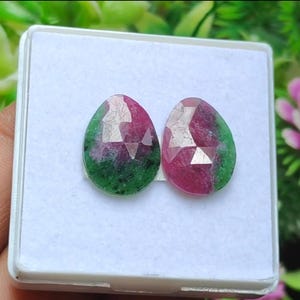 Può includere: Due gemme sfaccettate a forma di goccia. Ogni gemma presenta un vivace mix di colori: rosso intenso, verde smeraldo e sfaccettature chiare. Le gemme sono esposte in un contenitore quadrato bianco, mettendo in risalto le loro superfici lucide e le variazioni cromatiche naturali.