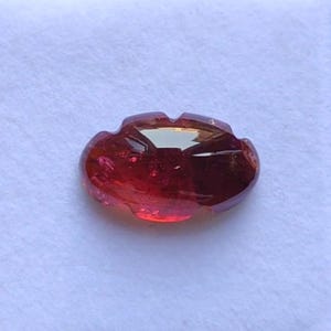 May include: A faceted, oval-shaped gemstone with a deep red hue. The gem has a flower-like design with rounded edges and a polished surface, reflecting light. The gemstone is set against a white background.