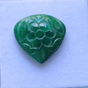 May include: A vivid green gemstone with a floral carving. The teardrop-shaped stone features a detailed carving of a flower and leaves. The polished surface reflects light, accentuating the intricate details of the floral design.