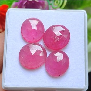 May include: Four faceted, oval-shaped, bright pink gemstones are displayed in a white rectangular container. The gems have a polished, reflective surface with multiple facets that catch the light, creating a sparkling effect. The gems are a vibrant shade of pink.
