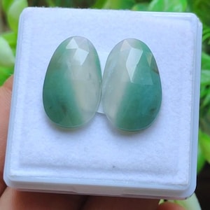 May include: Two oval gemstones with a faceted surface. Each gem has a gradient of white and green hues, creating a striking contrast. The stones are displayed in a white, rectangular box, showcasing their polished appearance.