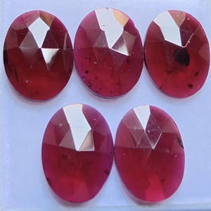 Può includere: Cinque pietre preziose rosse di forma ovale e sfaccettate, disposte in gruppo. Ogni pietra presenta una superficie lucida con molteplici sfaccettature triangolari che riflettono la luce, mostrando una profonda tonalità cremisi con piccole inclusioni scure.