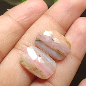 May include: Two faceted, oval-shaped gemstones. The stones display a mix of pink, white, and gold hues. The facets catch the light, creating a sparkling effect. The gemstones are likely for jewelry making.