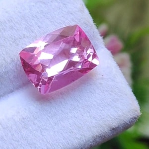 Könnte beinhalten: Ein facettierter, rechteckiger rosa Edelstein mit Brillantschliff. Der Edelstein hat einen leuchtenden Rosaton und reflektiert das Licht über seine vielen Facetten. Der Edelstein ist vor einem weißen Hintergrund platziert.