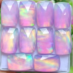 May include: A collection of eight faceted, rectangular gemstones with a pearlescent, iridescent quality. The stones display a range of colors, including pink, purple, and hints of yellow and green. The gems are arranged in a grid pattern.