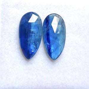 May include: Two pear-shaped blue kyanite gemstones with faceted tops. The stones exhibit a deep royal blue colour with visible internal inclusions and natural striations throughout the surface, reflecting light from their polished upper facets.