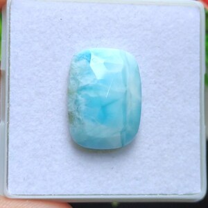 May include: A rectangular, light blue gemstone with a polished, faceted surface. The stone has a cloudy, marbled appearance with shades of white and turquoise. The gemstone is set against a white background.