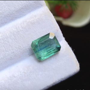 May include: A rectangular emerald-cut gemstone with a vibrant green hue and visible internal inclusions. The stone features faceted edges that reflect light, showcasing a translucent quality and a polished surface, resting on a white fabric.
