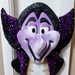May include: A whimsical, framed wall decoration featuring a cartoon vampire face. The vampire has a purple velvet face, black glitter hair, and purple sequined wings. The vampire's outfit includes a white shirt with gold trim.
