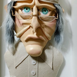 Op de afbeelding: Een witte viltpop met een humeurige uitdrukking, die een witte slaapmuts en een bril draagt. De pop heeft een grote neus en een lange witte baard. De pop draagt een beige shirt met drie witte knopen.