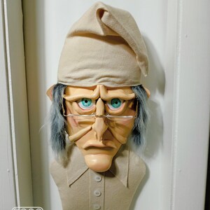 Op de afbeelding: Een beige vilten sculptuur van een mensenhoofd met een slaapmuts, blauwe ogen, een grote neus en een beige shirt met knopen. Het beeld is bevestigd aan een witte muur.