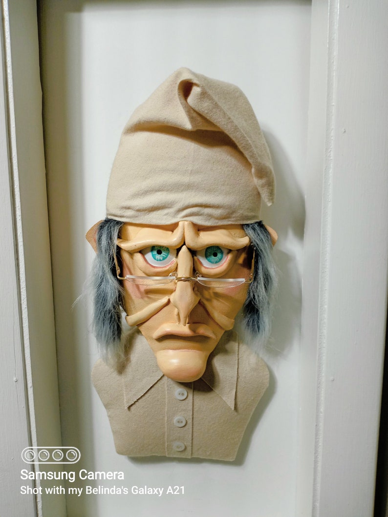 Op de afbeelding: Een beige vilten sculptuur van een mensenhoofd met een slaapmuts, blauwe ogen, een grote neus en een beige shirt met knopen. Het beeld is bevestigd aan een witte muur.