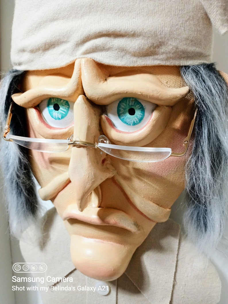 Op de afbeelding: Een close-up van het gezicht van een pop met een witte hoed, grijs haar en een serieuze uitdrukking. De pop draagt een bril met gouden accenten en heeft blauwe ogen met turquoise pupillen.