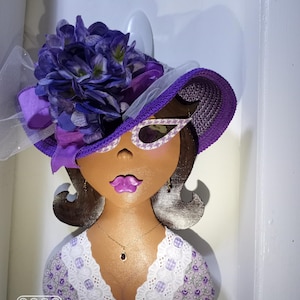 May include: A purple and white hat with a flower on top, a brown face with purple sunglasses, and a purple and white patterned shirt. The face has a pink mouth and brown eyes.