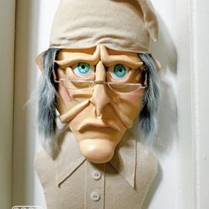 Op de afbeelding: Een beige stoffen sculptuur van een persoon die een slaapmuts en een bril met blauwe ogen draagt. De sculptuur heeft een droevige uitdrukking en draagt een beige shirt met vier witte knopen.