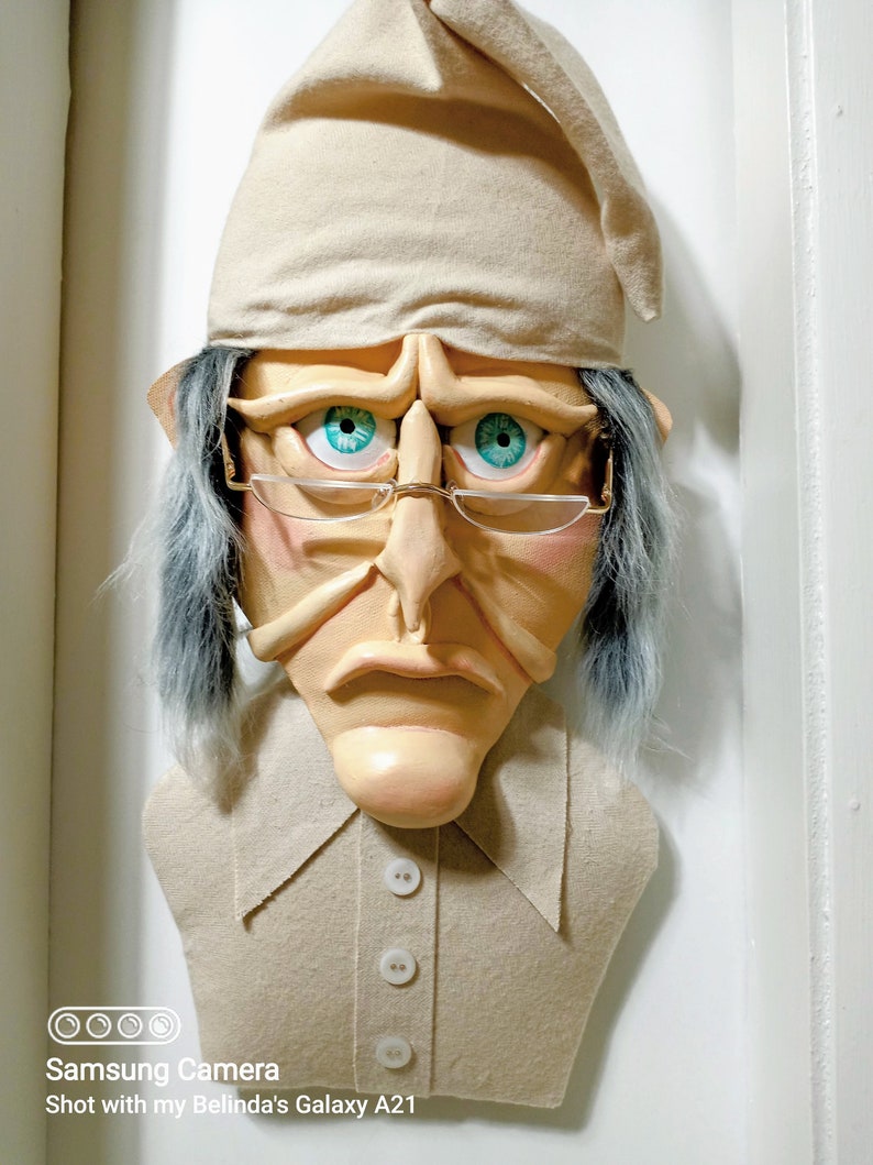 Op de afbeelding: Een beige stoffen sculptuur van een persoon die een slaapmuts en een bril met blauwe ogen draagt. De sculptuur heeft een droevige uitdrukking en draagt een beige shirt met vier witte knopen.