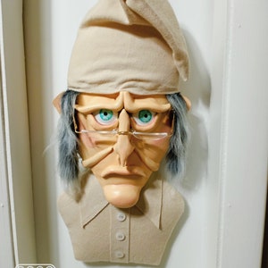 Op de afbeelding: Een beige vilt sculptuur van een persoon die een beige slaapmuts, een bril en een beige shirt met knopen draagt. Het beeld heeft een chagrijnige uitdrukking en blauwe ogen.