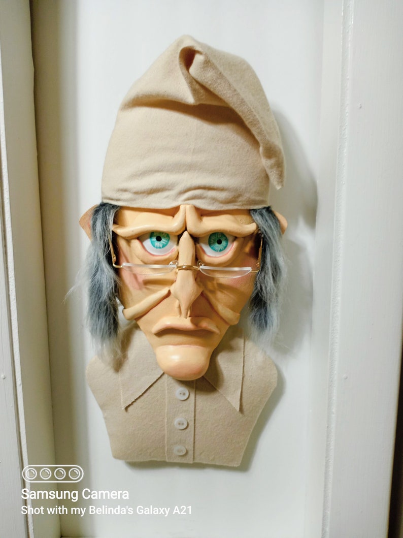 Op de afbeelding: Een beige vilt sculptuur van een persoon die een beige slaapmuts, een bril en een beige shirt met knopen draagt. Het beeld heeft een chagrijnige uitdrukking en blauwe ogen.