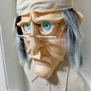 Op de afbeelding: Een witte stoffen buste van een persoon met een witte slaapmuts, grijs haar en een serieuze uitdrukking. De buste draagt een wit shirt met knopen. De persoon heeft blauwe ogen en draagt een bril.