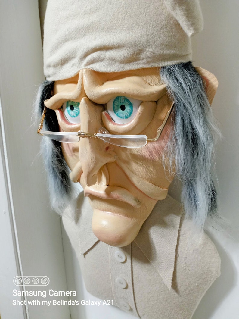 Op de afbeelding: Een witte stoffen buste van een persoon met een witte slaapmuts, grijs haar en een serieuze uitdrukking. De buste draagt een wit shirt met knopen. De persoon heeft blauwe ogen en draagt een bril.