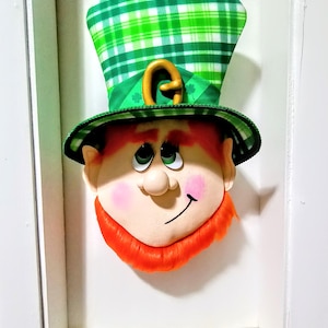 May include: A green and white plaid leprechaun hat with a gold buckle and a red beard. The leprechaun's face is painted with a smile and big eyes.