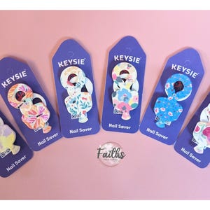Può includere: Sei salvanchiodi colorati con motivi floreali su uno sfondo viola. Ogni salvanchiodo ha la scritta "KEYSIE" e "Nail Saver" stampata su di esso.