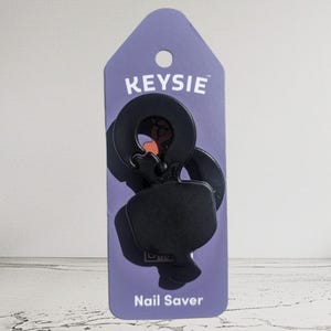 Può includere: Un accessorio portachiavi KEYSIE Nail Saver nero su un'etichetta viola. Il portachiavi ha un design unico con una parte superiore circolare e una parte inferiore curva. L'etichetta ha il nome del marchio "KEYSIE" e il nome del prodotto "Nail Saver" stampati.