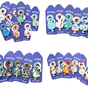 Può includere: Assortimento di anelli salva-unghie colorati con vari motivi, tra cui disegni floreali, animalier e geometrici. Ogni anello è etichettato con il nome del marchio "KEYSIE" e le parole "Nail Saver".