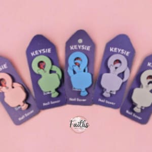 Puede incluir: Cinco protectores de uñas en varios colores, incluyendo rosa, verde, azul y gris, presentados en etiquetas moradas. Cada etiqueta lleva la inscripción "KEYSIE" y "Nail Saver". Fondo rosa claro.