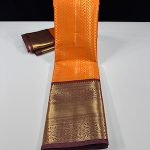May include: A vibrant orange saree featuring intricate gold woven patterns throughout the main body. The garment includes a wide, deep purple border adorned with elaborate gold zari floral motifs and geometric designs along the bottom edge.